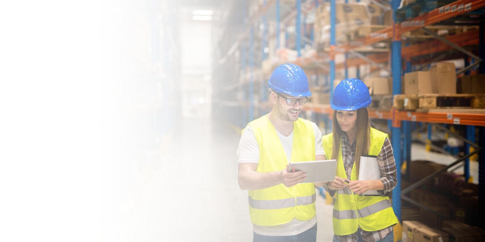 Dos trabajadores de almacén con cascos y chalecos de seguridad, revisando datos en una tablet, con estanterías de almacén de fondo.