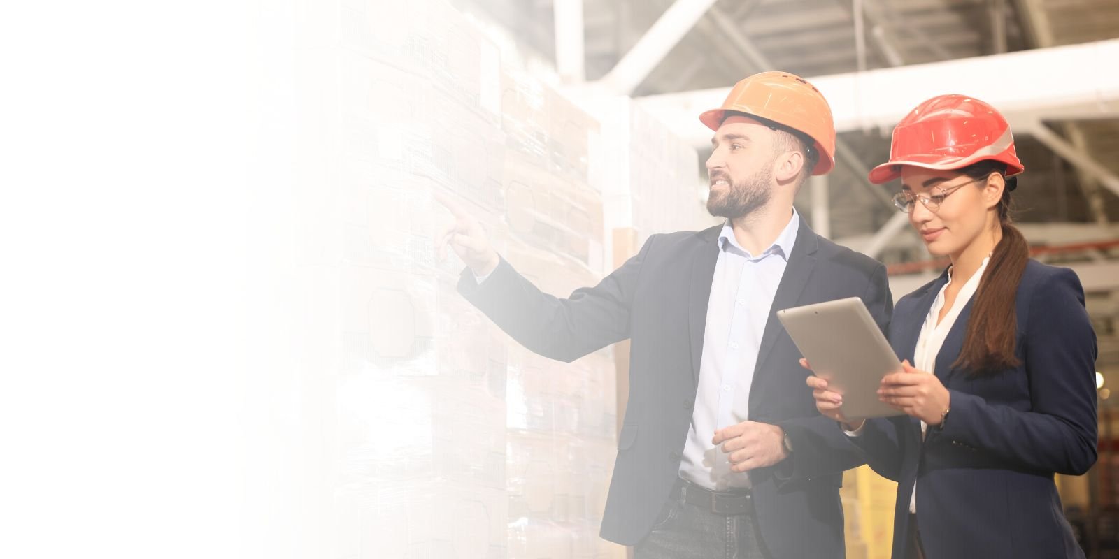 Un supervisor de almacén y una coordinadora de logística, ambos con cascos de seguridad, revisan productos y documentación en una tablet en un pasillo de almacén.